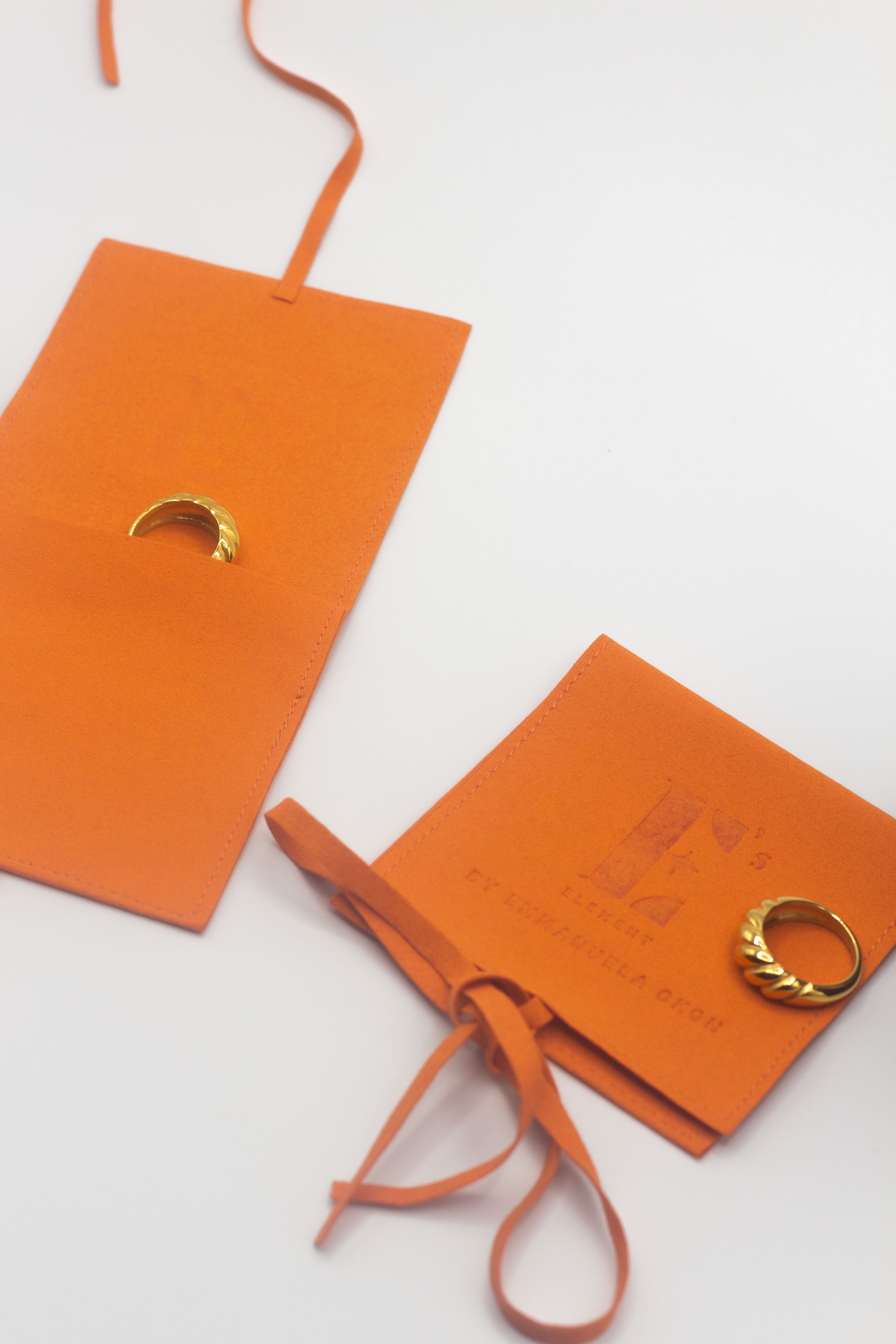 On the left is an 18k gold ring resting inside of an orange leather microfiber pouch. On the right is an 18k gold ring resting on an orange microfiber pouch with the E's Element logo engraved. Microfiber Jewelry Pouch by E's Element.