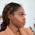 Woman in a light brown suit wearing a 18k gold stainless steel earring. The earring have a cross charm attached to it. Cross Cubic Zirconia Huggies by E's Element.