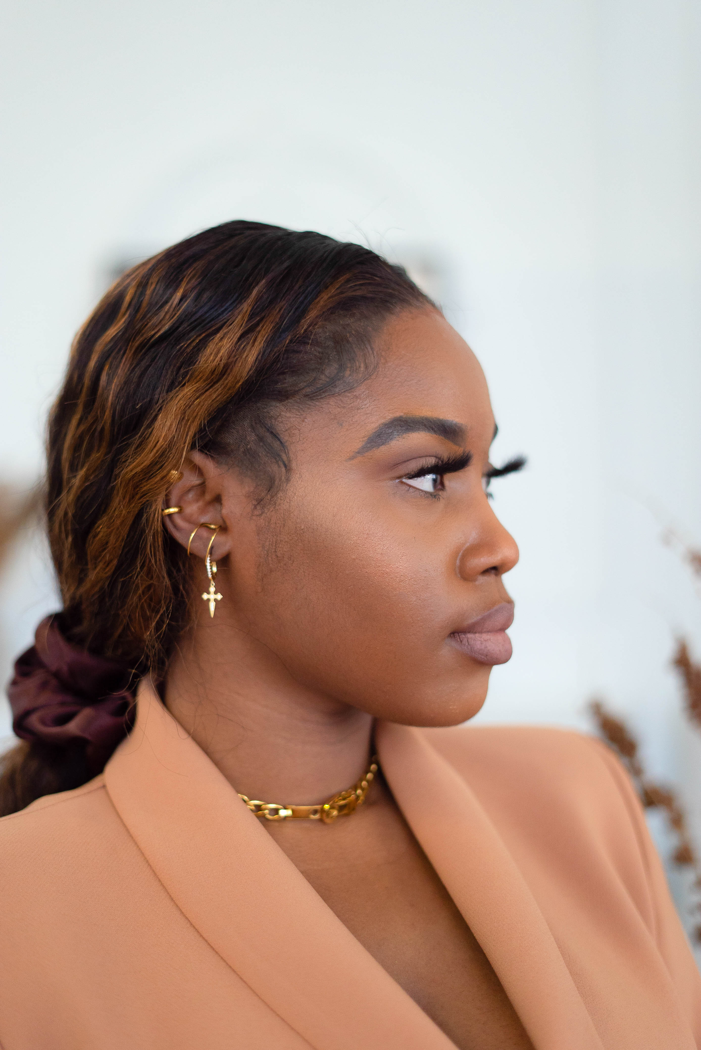 Woman in a light brown suit wearing a 18k gold stainless steel earring. The earring have a cross charm attached to it. Cross Cubic Zirconia Huggies by E's Element.