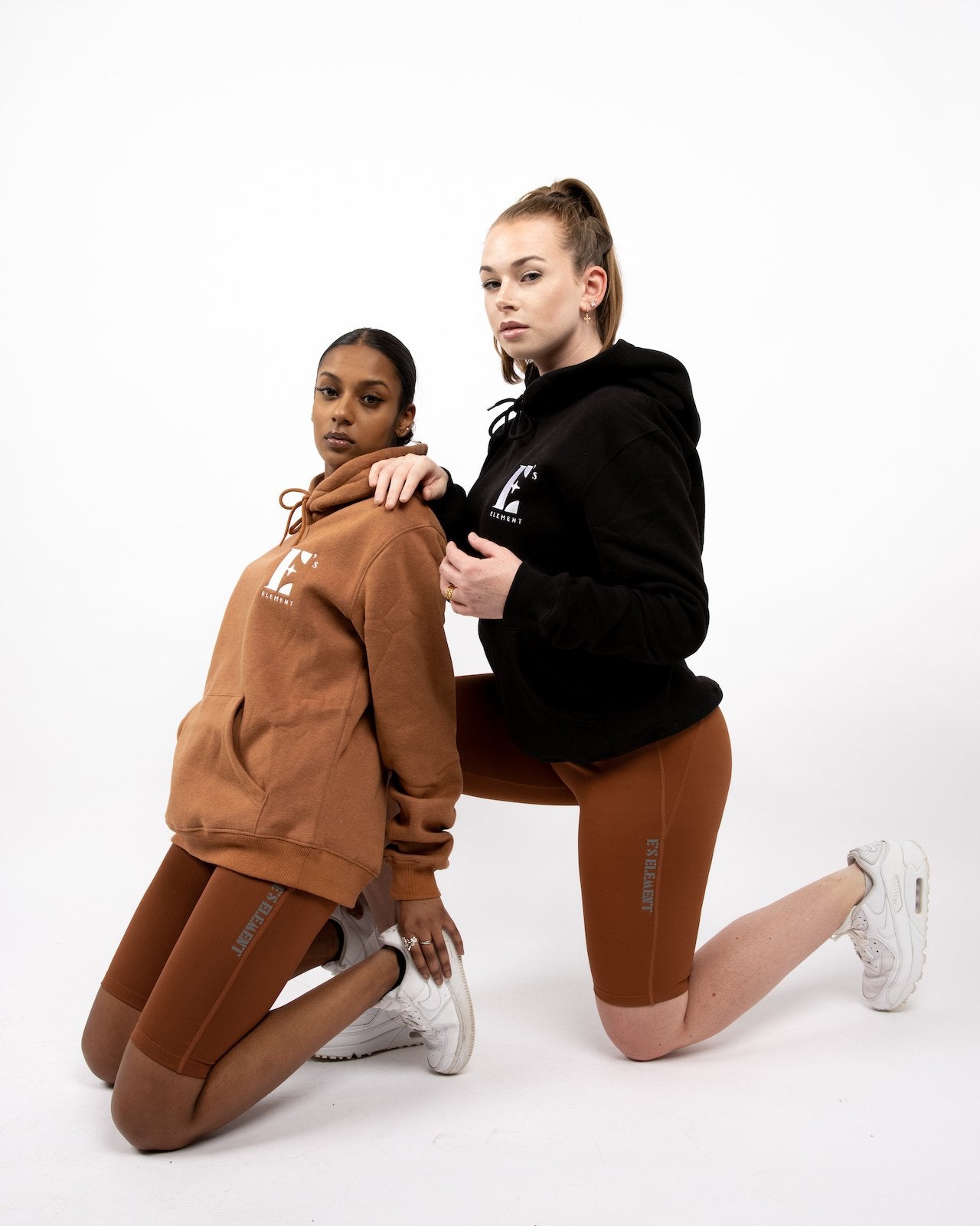 The model on the left is wearing a light brown hoodie. The model on the right is wearing a black hoodie. Both of the models are wearing brown biker shorts and white sneakers. The Ella Shaping Cycling Shorts (Petite Size) by E's Element.