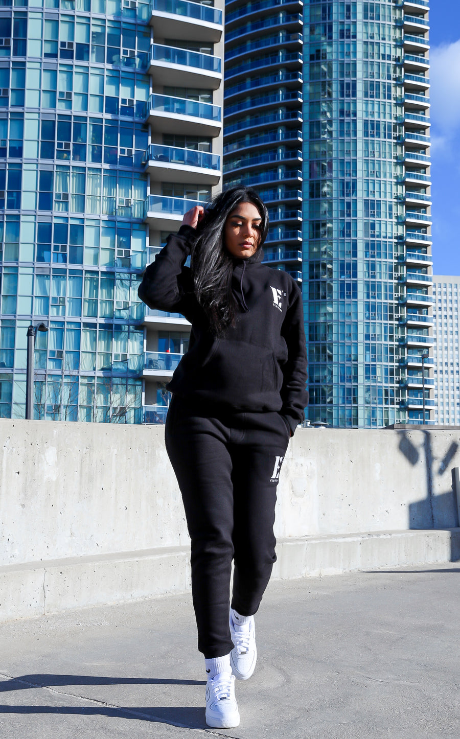Model wearing black sweat suit. The hoodie and sweat pants has the E's Element logo imprinted on it in white. The model is also wearing white sneakers. E's Element Essential Smoky Black Sweatsuit Set by E's Element.