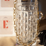 18k gold chain necklace with a rope pattern. The necklace is hanging on the edge of a glass cup. Behind the cup are two white boxes stacked on each other with the E's Element logo in red. Rope Link Signature Chain by E's Element.