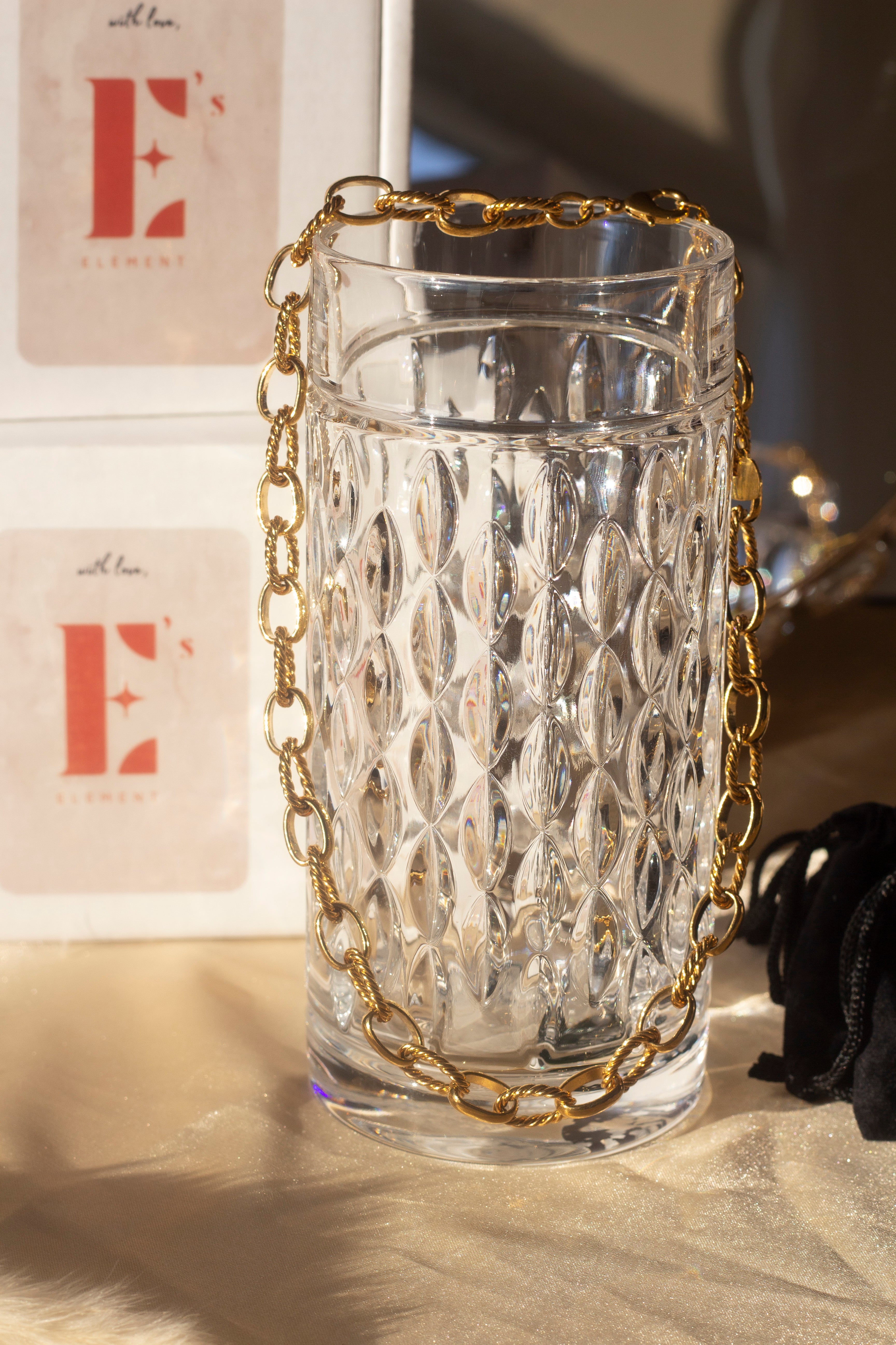 18k gold chain necklace with a rope pattern. The necklace is hanging on the edge of a glass cup. Behind the cup are two white boxes stacked on each other with the E's Element logo in red. Rope Link Signature Chain by E's Element.
