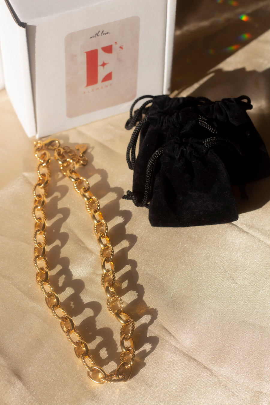 18k gold chain necklace with a rope pattern. The chain is placed on a beige cloth. On the top left is a box with the E's Element logo in red. On the right are closed black pouches. Rope Link Signature Chain by E's Element.