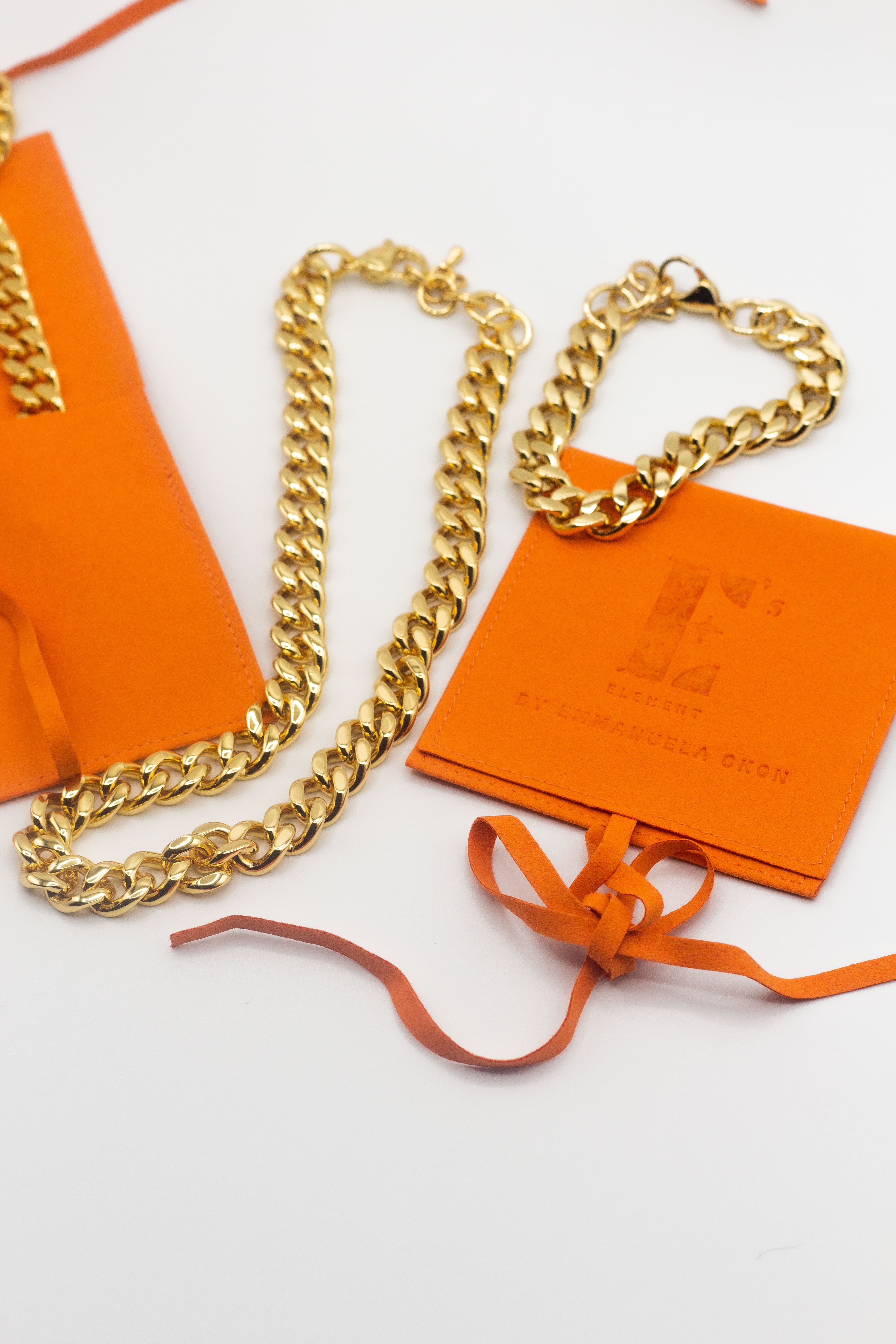 18k gold chain necklace in the middle. On the right is an 18k gold chain bracelet placed on an orange leather pouch. The Emmanuela Set in Gold by E's Element.