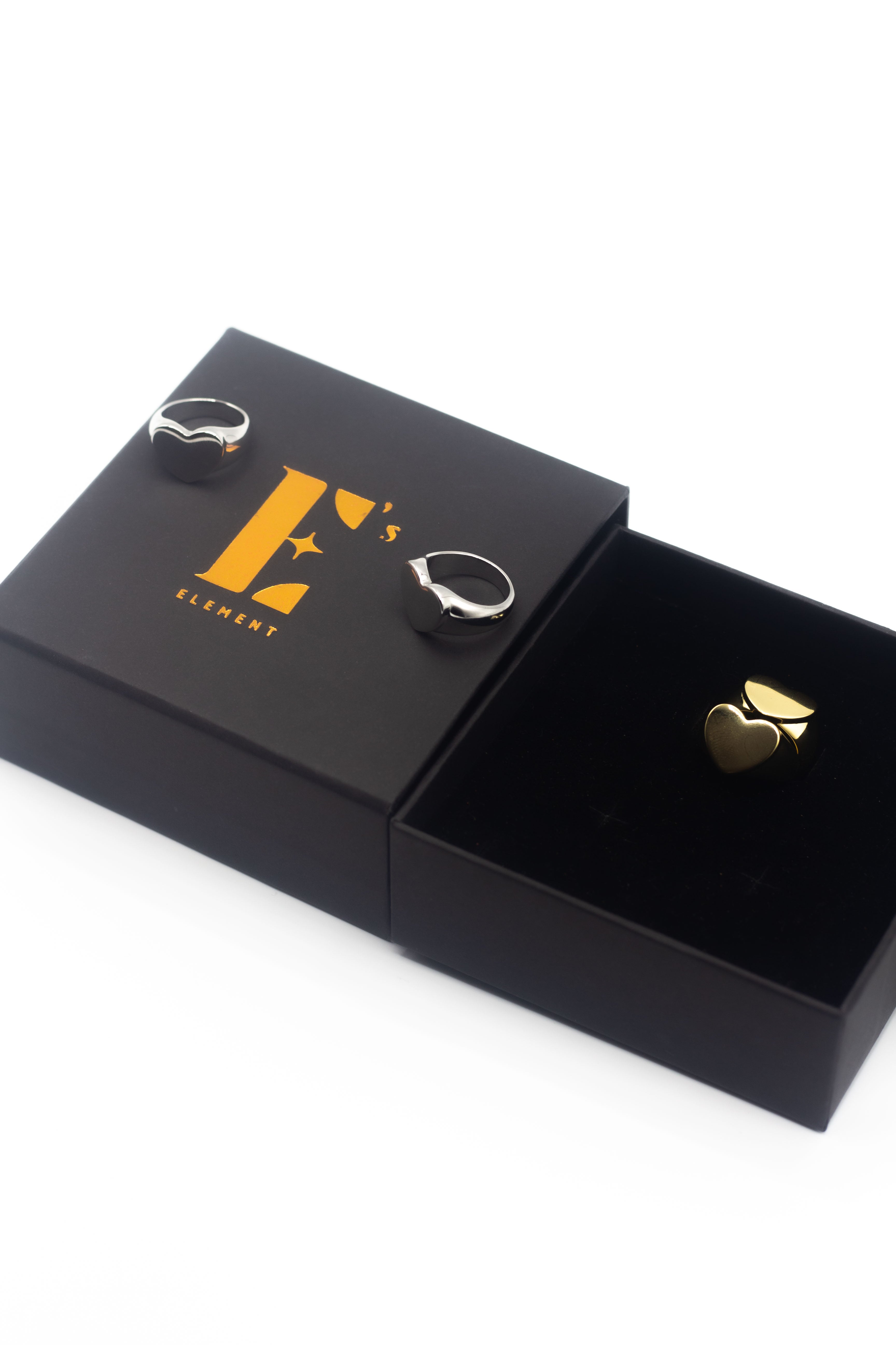On the left there is a black box with the E's Element logo imprinted in yellow. Resting on top of the box are two 18k silver rings with heart signets. On the right is an opened box with an 18k gold ring with a heart shaped signet. Ellina Heart Signet Ring by E's Element.