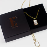 18k gold necklace with a white rectangular charm placed in its container. On the left is the cap for the container with the E's Element imprinted in gold. The Infinity Zircon Necklace by E's Element.
