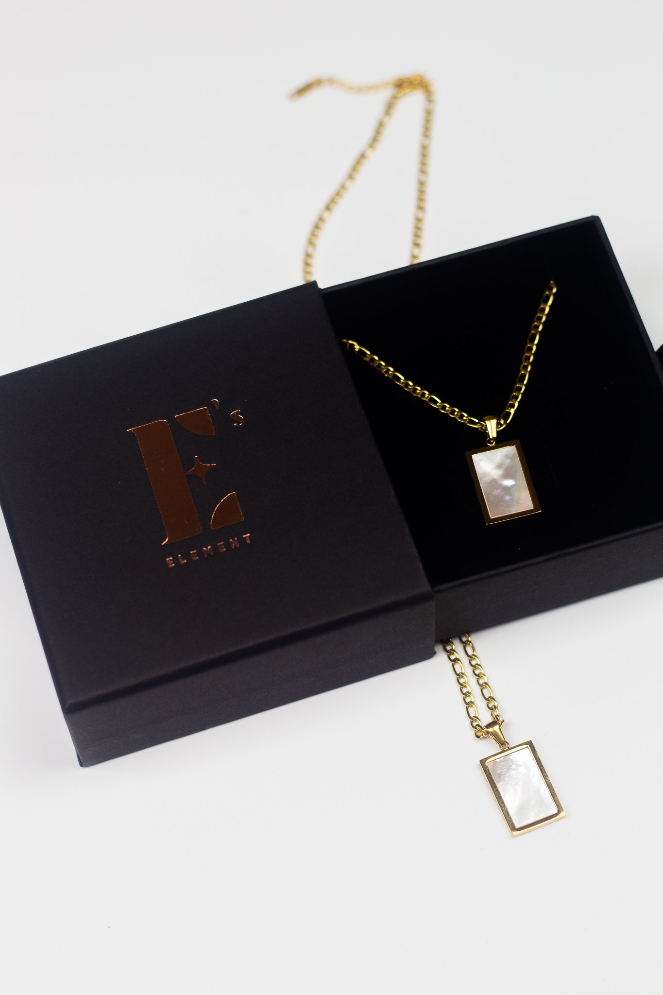 18k gold necklace with a white rectangular charm placed in its container. On the left is the cap for the container with the E's Element imprinted in gold. The Infinity Zircon Necklace by E's Element.