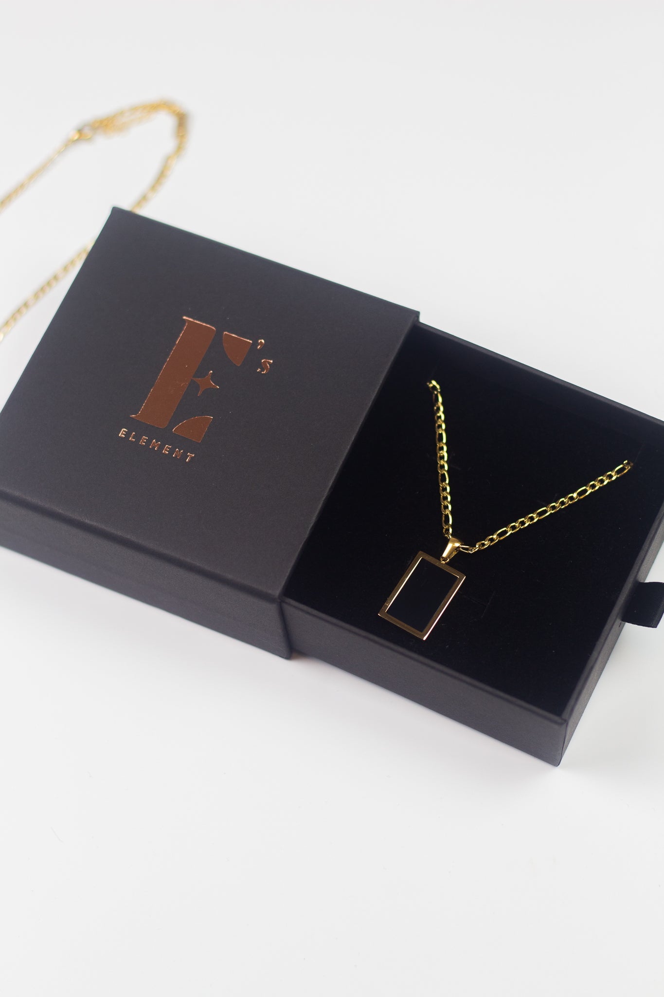 18k gold necklace with a black rectangular charm placed in its container. On the left is the cap for the container with the E's Element imprinted in gold. The Infinity Zircon Necklace by E's Element. 