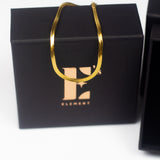 18K gold snake chain anklet resting on top of its container. E's Element is labelled on the container in gold. The anklet is named "The Steph" Herringbone Anklet by E's Element