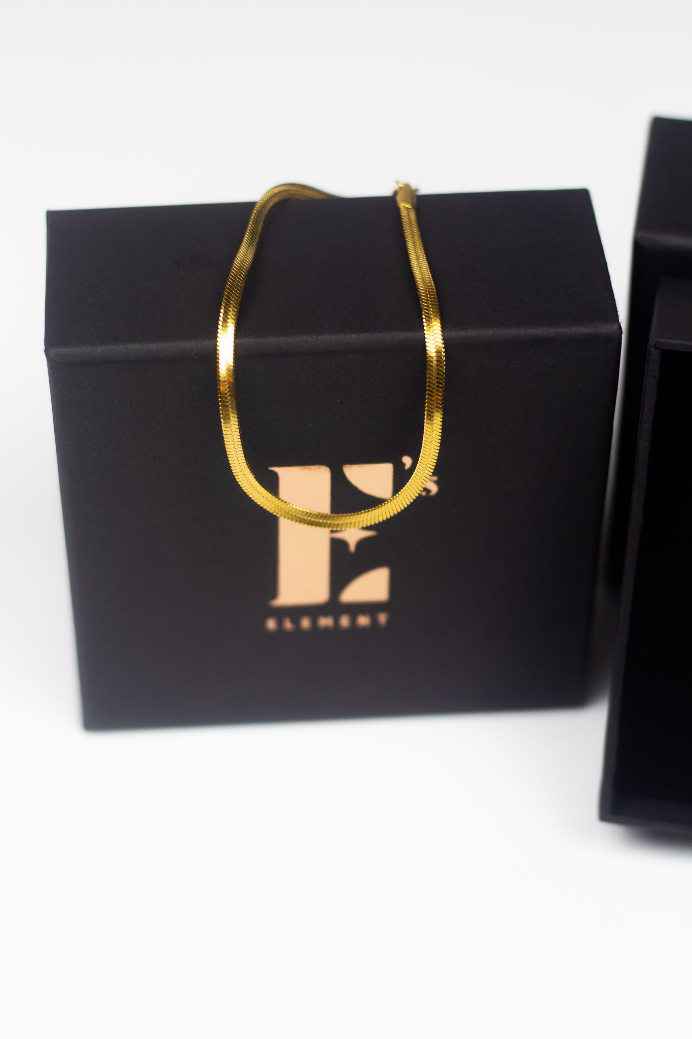 18K gold snake chain anklet resting on top of its container. E's Element is labelled on the container in gold. The anklet is named "The Steph" Herringbone Anklet by E's Element