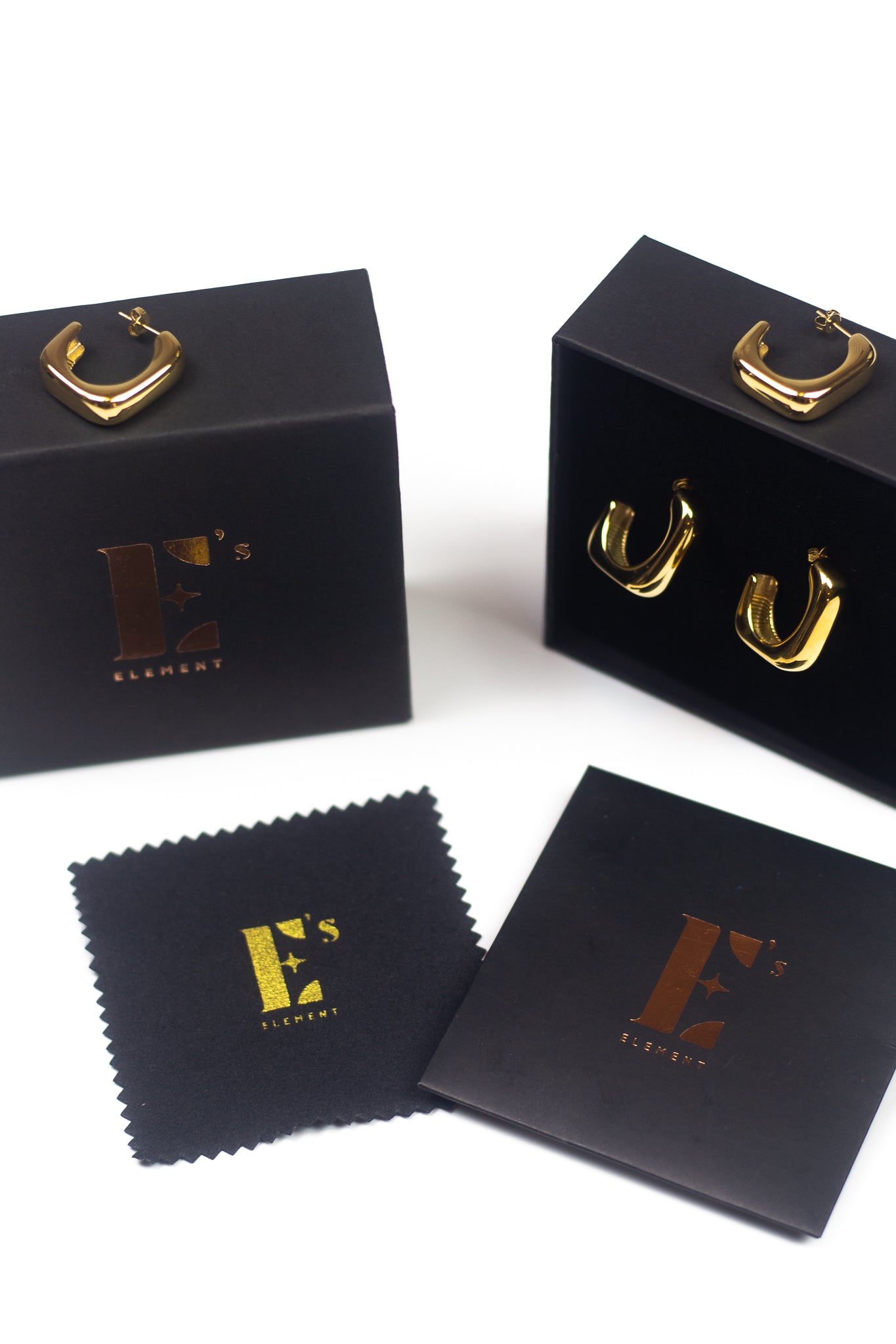 18k gold stainless steel hoop earrings resting on top of its container. There is a polishing cloth and business card with E's Element logo printed in gold underneath. Angel Hoops by E's Element.