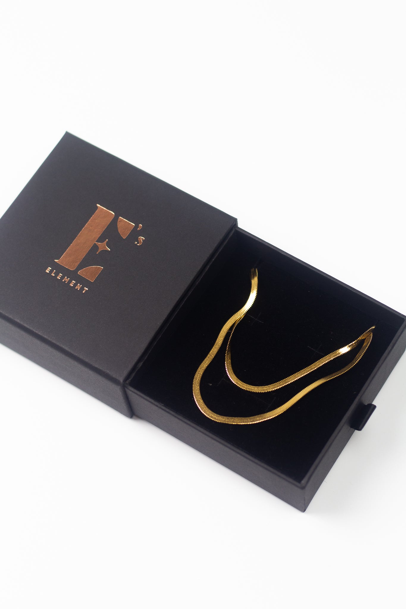 18K gold herringbone chain necklace resting in its container beside the cap of the container labelled with E's Element logo. Named "The Steph" Herringbone Chain Necklace by E's Element.