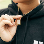 Model in a black hoodie wearing 18k gold necklace with a white rectangular charm. The hoodie has the E's Element logo in white. The Infinity Zircon Necklace by E's Element.