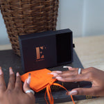 Orange microfiber leather pouch resting on a black book. Behind the pouch is a black E's Element box and E's Element logo imprinted in gold. Microfiber Jewelry Pouch by E's Element.