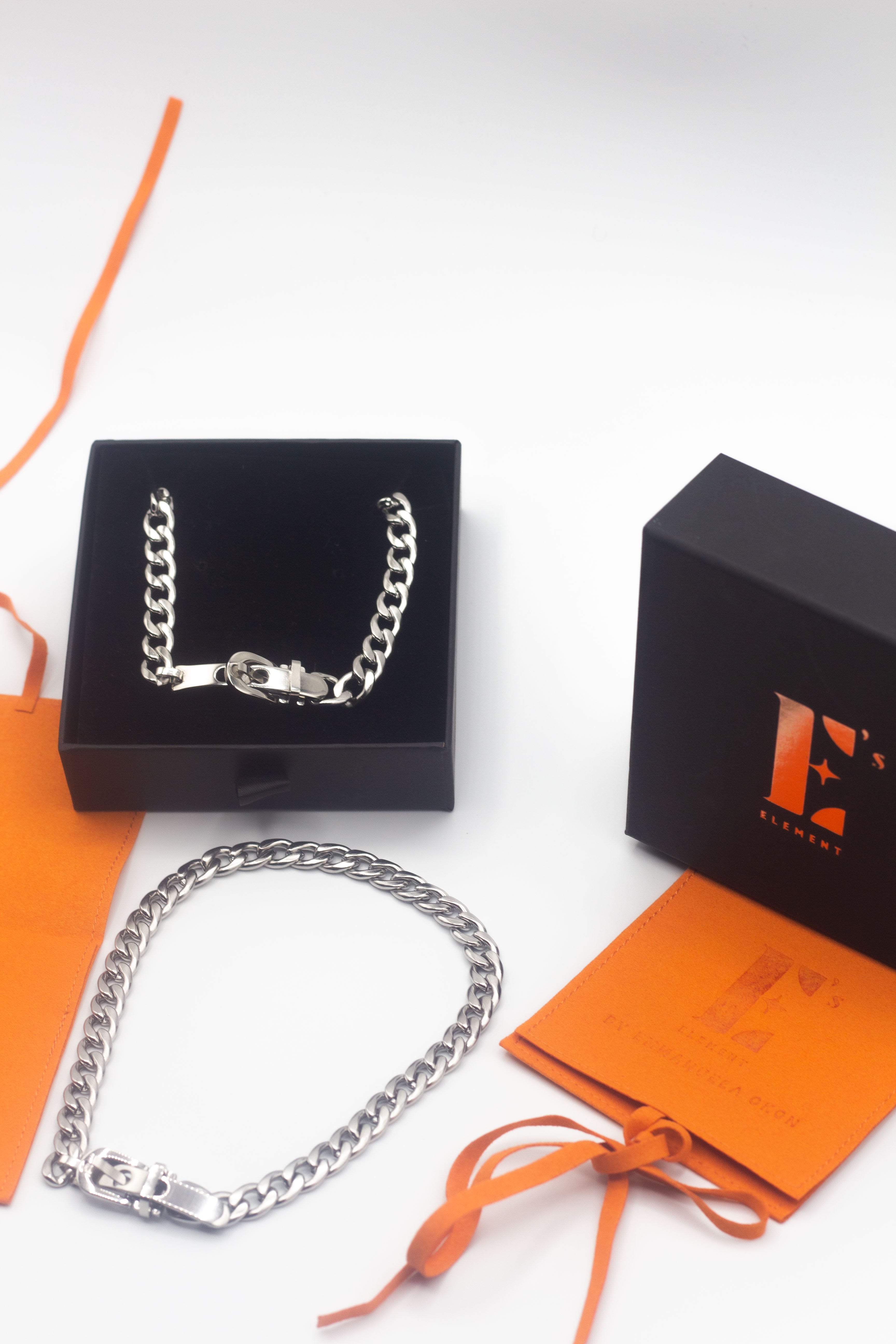 18k silver stainless steel buckle choker resting in its container. There is an orange leather packaging on the right and a lid with the E's Element logo imprinted. There is another silver buckle choker placed under the container with the choker. Ella Buckle Choker by E's Element.