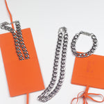 Stainless steel chain necklace and bracelet placed on top of orange leather pouches. The Emmanuela Set in Steel by E's Element.