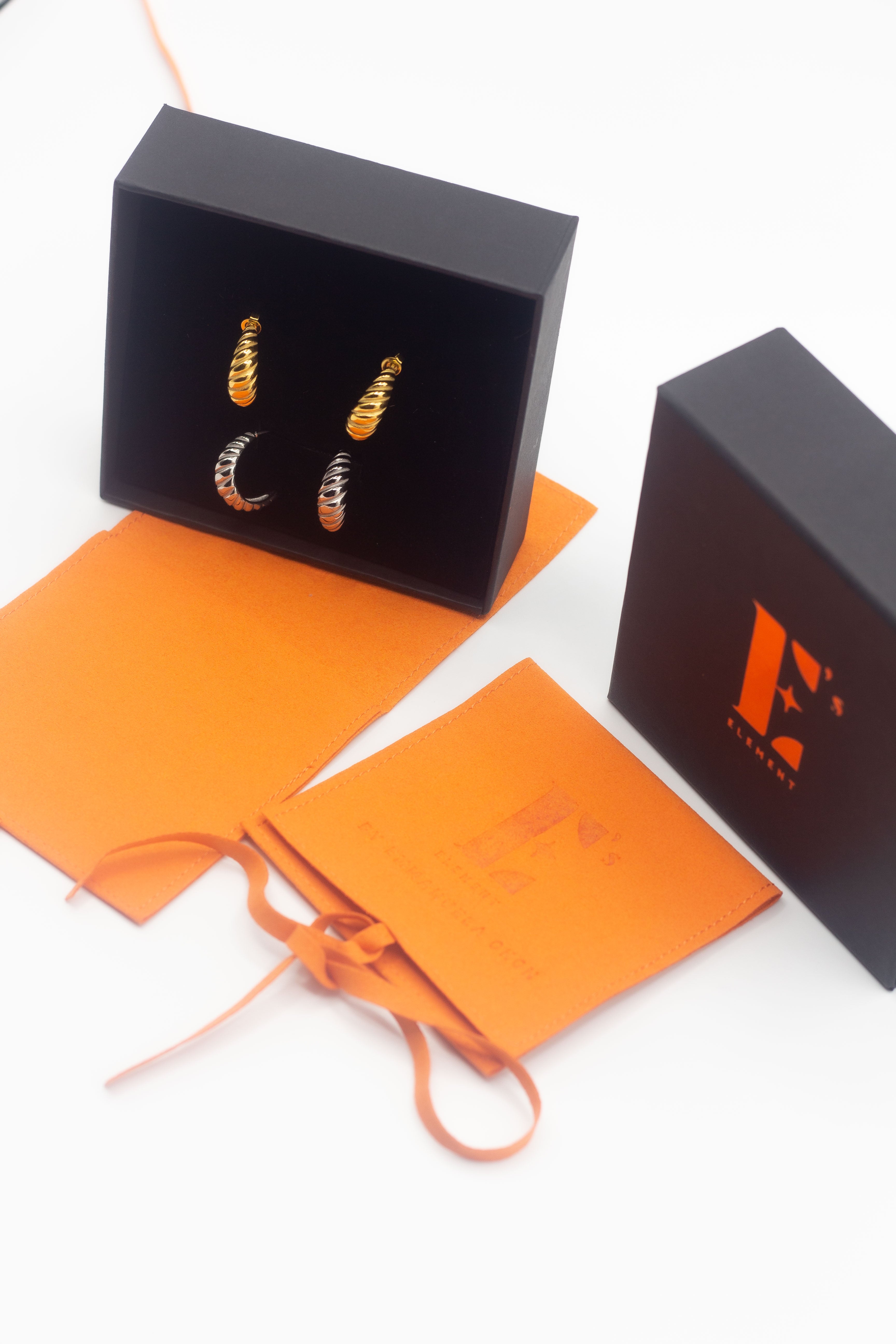 18k gold and silver earrings placed in a black opened container on the left. On the right is the cap for the container with the E's Element logo in gold. Under the box and cap are orange leather pouches. Thick Croissant Stud Earrings - E's Element.