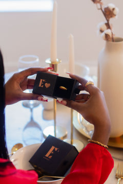 Model in a red jacket holding a black container with the E's Element logo and a heart in gold. Inside the container is an 18k gold ring. Valentine's Day Ring Box (Add-On) - E's Element.