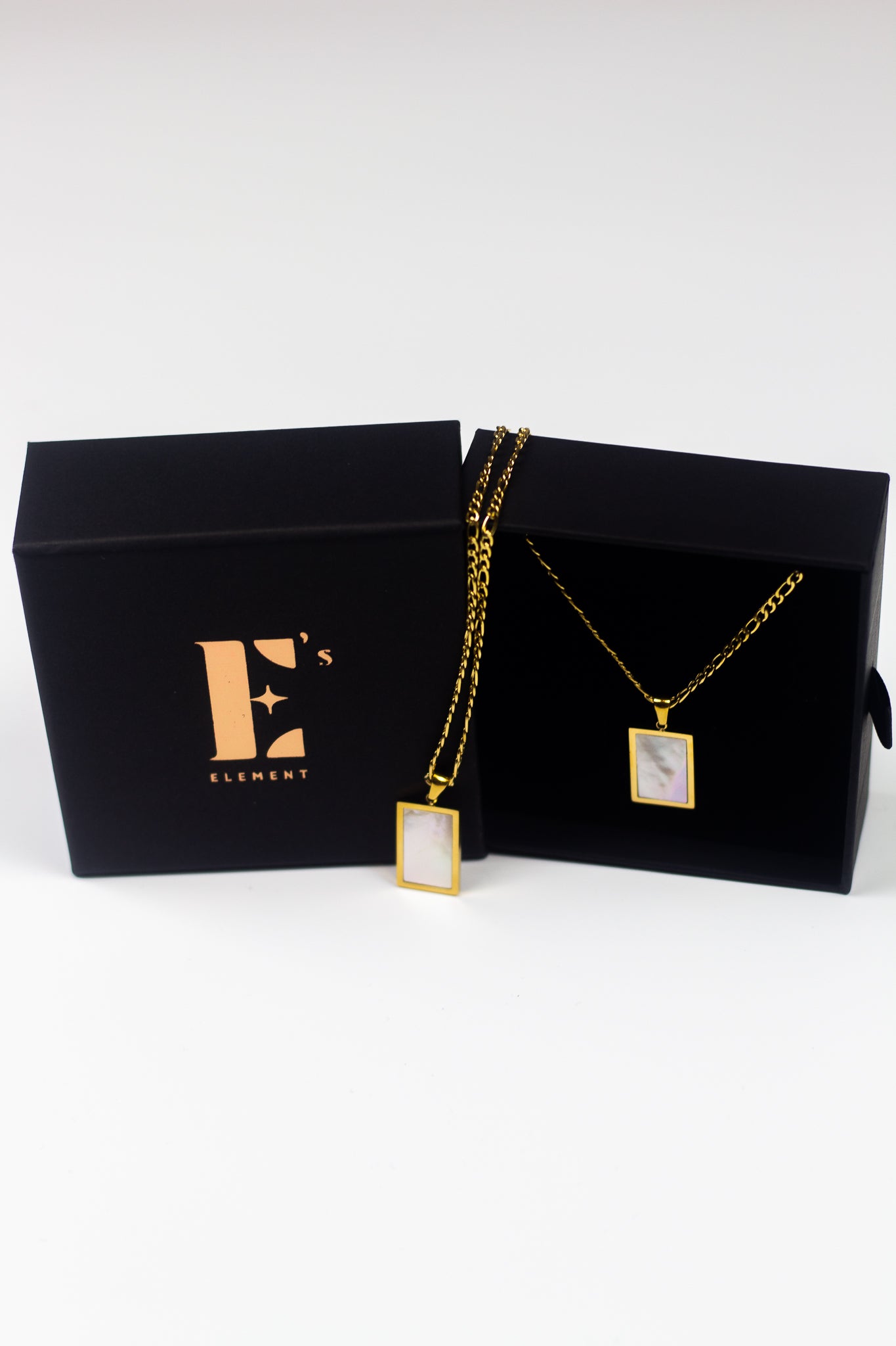 18k gold necklace with a white rectangular charm placed in its container. On the left is the cap for the container with the E's Element imprinted in gold. The Infinity Zircon Necklace by E's Element. 
