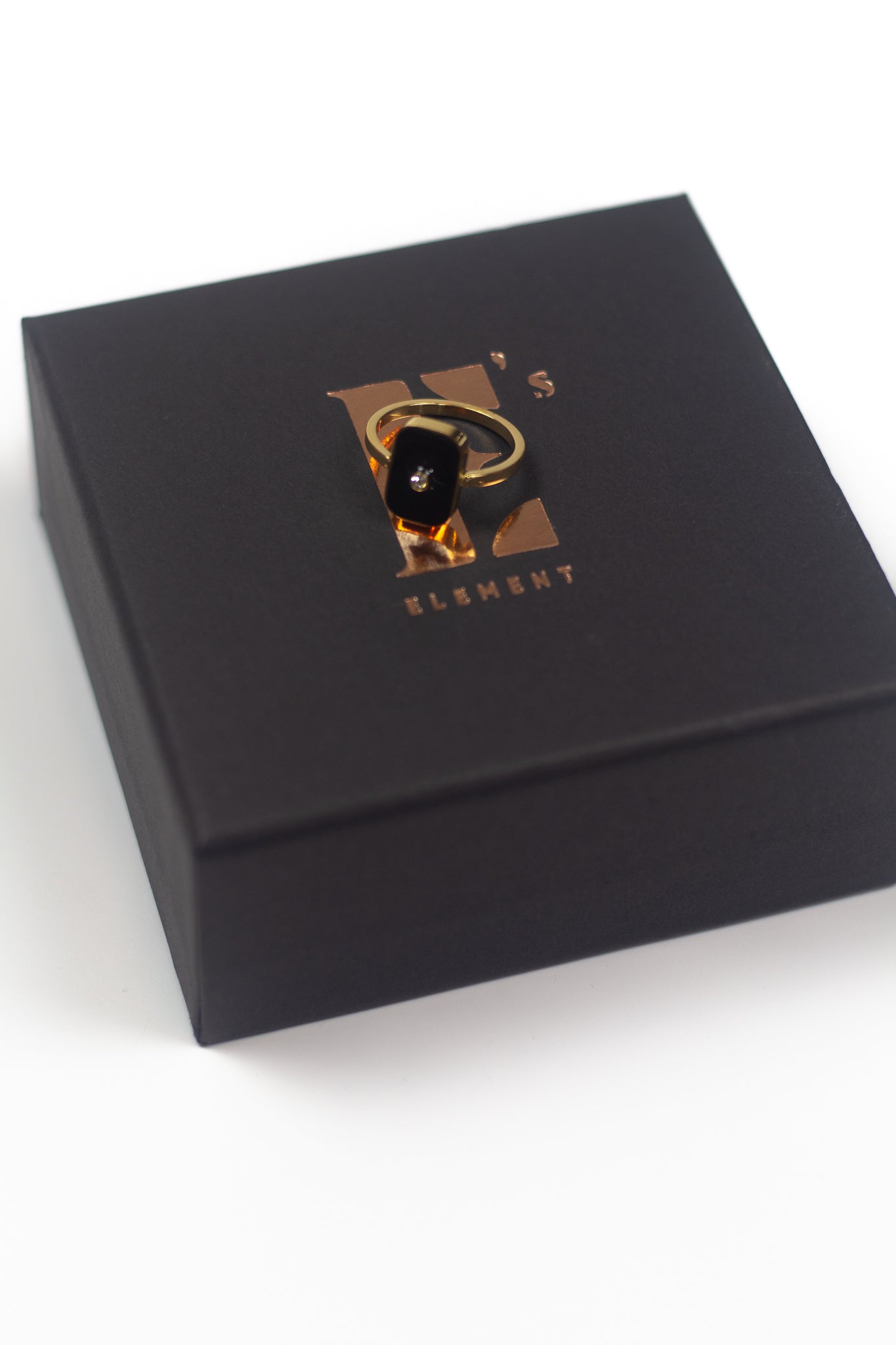 18k gold ring resing on a black container with the E's Element logo in gold. The ring has a zircon stone in the middle of a flat rectangular surface in black. Infinity Zircon Ring by E's Element.